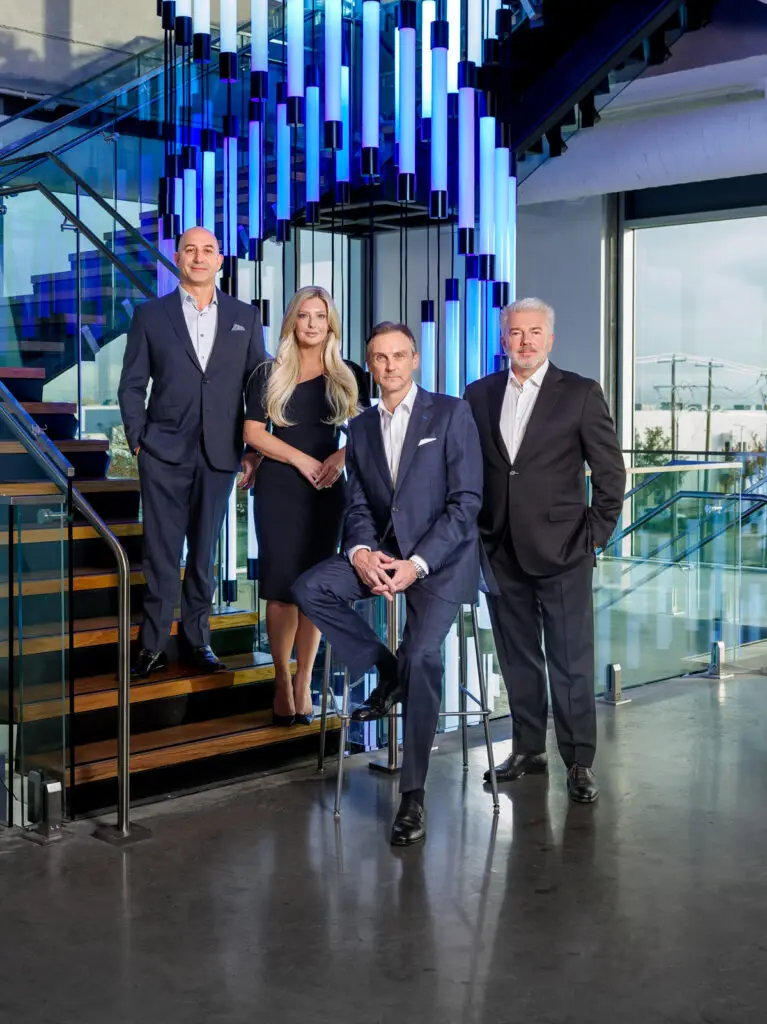 Four Professionally Dressed Individuals Posing in a Modern Office Lobby with Blue Lit Hanging Tubes in the Background