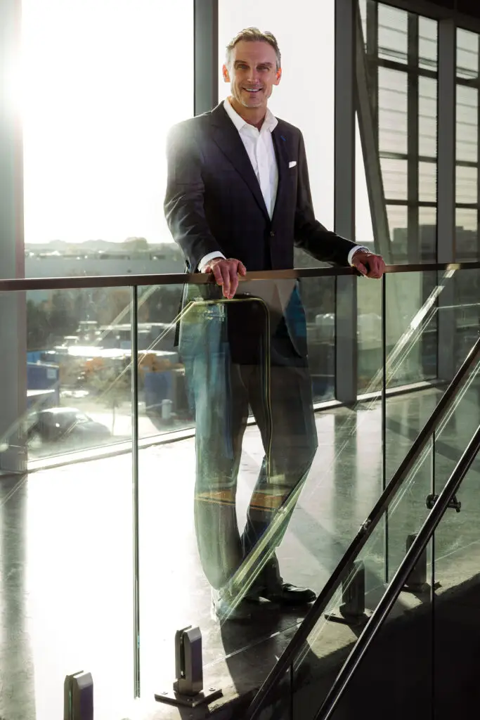 Smiling Businessman in a Dark Suit Leaning on a Glass Railing in a Bright Modern Office Atrium