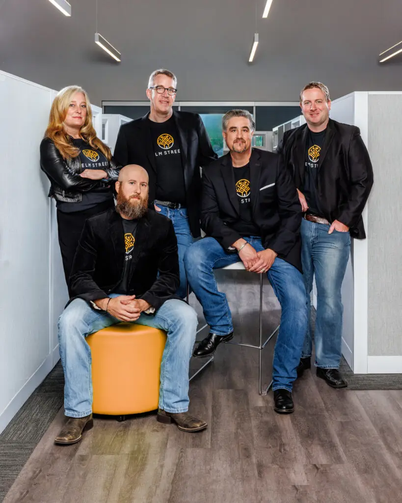 Five Colleagues Pose in a Modern Office Wearing Matching Black Jackets and Elm Street T Shirts One Man Sits on an Orange Ottoman While Four Stand Around Him
