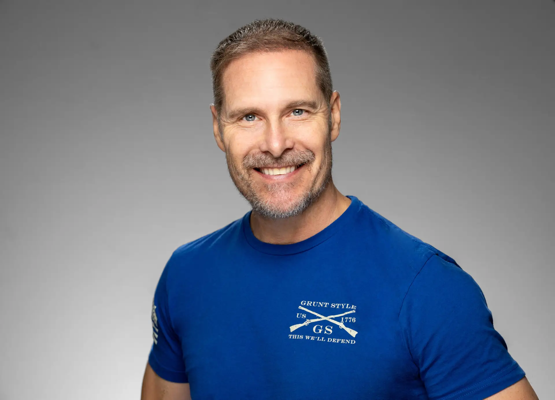 Smiling Man with Short Gray Beard Wearing a Blue Grunt Style Shirt Against a Gray Studio Backdrop