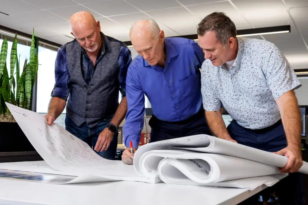 Three Men Examine Large Architectural Blueprints on a Drafting Table Marking Details with a Red Pen and Pencil in a Bright Office