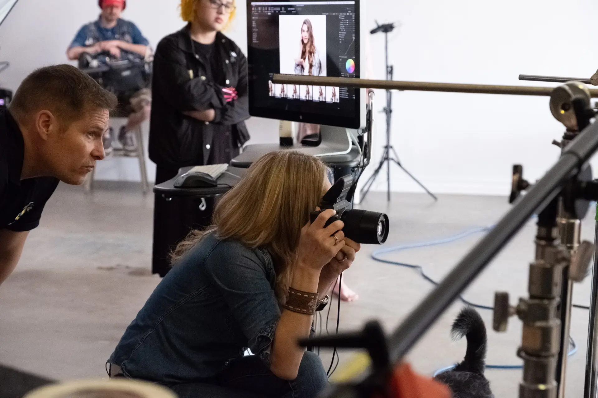 Photographer Kneels with a Camera While a Model is Displayed on a Monitor in a Photography Studio with Crew and Gear in the Background