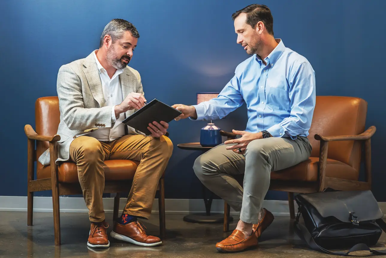 corporate-meeting-two-men - Kelly Williams Photography - Kelly Williams Photography Two Men in a Modern Office Sit on Leather Chairs One Showing a Tablet to the Other