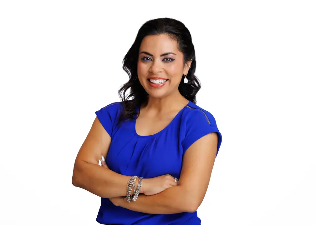 Smiling woman with dark hair in a blue blouse, arms crossed against a white background.