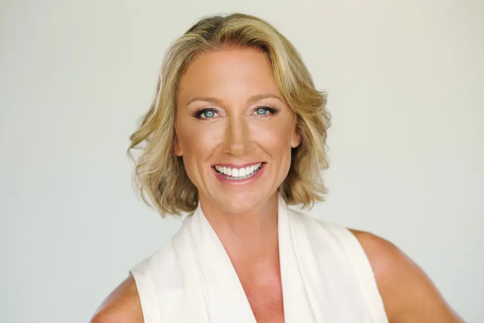 Close-up portrait of a smiling blonde woman with blue eyes in a white sleeveless top against a light neutral backdrop.