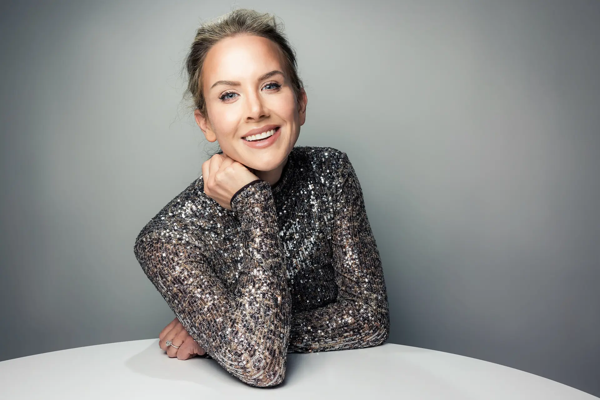 Smiling woman wearing a glittery sequined top, leaning on a white table with her chin resting on her hand.