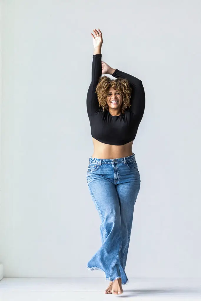 Woman with Curly Hair in a Black Crop Top and Jeans Dancing Barefoot Against a Light Gray Wall Arms Raised in a Stretch Pose