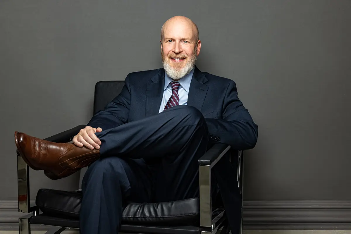 Smiling bald man with a white beard in a navy suit sits with legs crossed in a black leather chair in a neutral room.