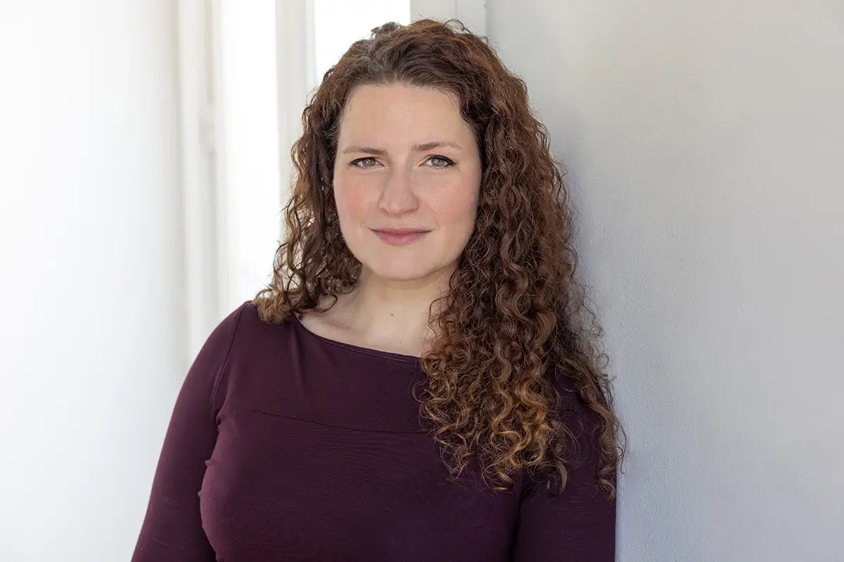 Portrait of a woman with curly brown hair in a maroon top, standing against a light wall.