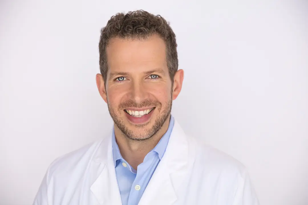 Smiling man in a white lab coat and light blue shirt against a plain white background, portrait-style.