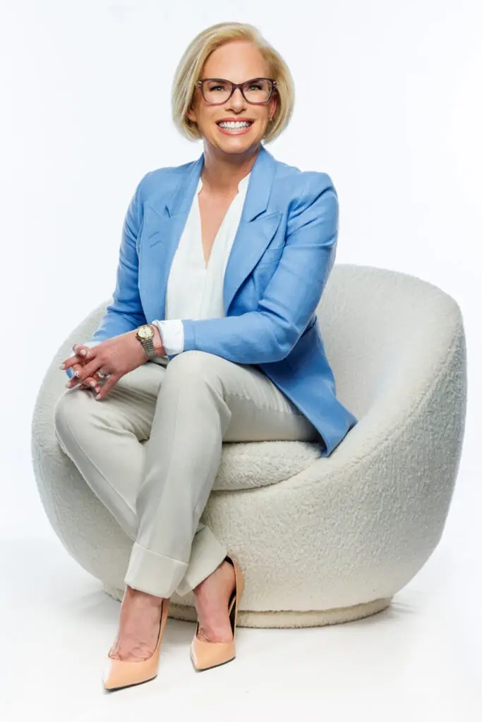 Smiling Woman in a Light Blue Blazer and Beige Pants Sits in a Plush Round Chair Posing for a Professional Portrait