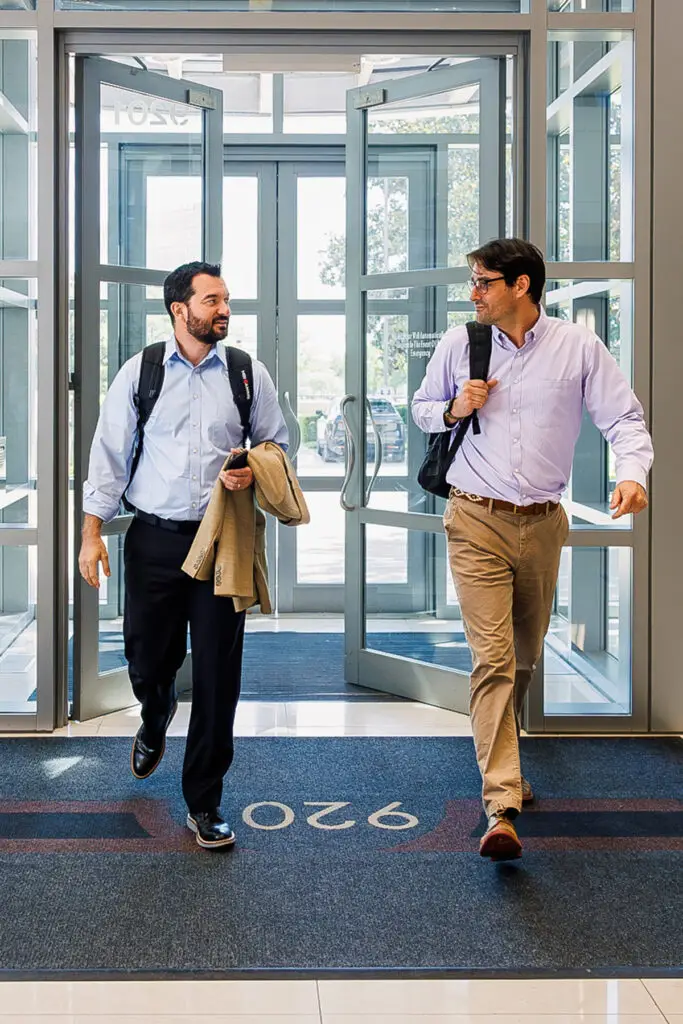 Two Businessmen Exit Through Glass Doors in a Modern Lobby Carrying Bags One with a Coat Draped over His Arm