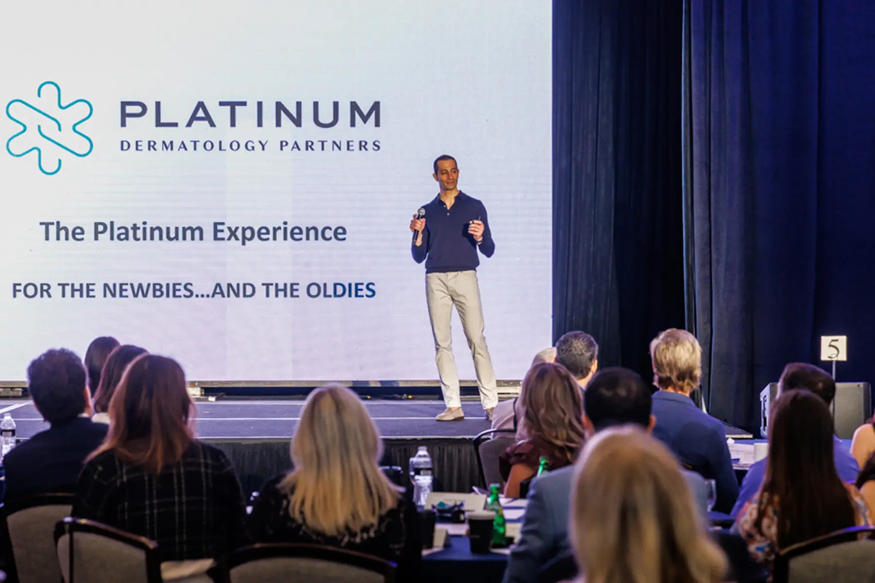 Male Presenter on a Stage Addressing a Seated Audience Behind Him is a Large Screen with Platinum Dermatology Partners and the Platinum Experience Text
