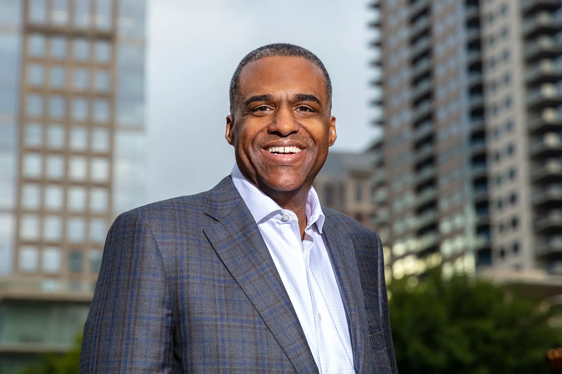 Smiling Man in a Blue Gray Plaid Suit Jacket Standing Outdoors in a City with Tall Buildings Behind Him