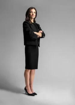 full length photo of beautiful dark haired executive woman wearing a black dress suit with arms folded and serious expression standing on a plain grey backdrop